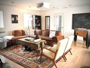 Un salon avec un canapé en cuir marron, un fauteuil blanc, une table basse, une bibliothèque et une table de baby-foot.