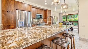 A modern kitchen with granite countertops, stainless steel appliances, wooden cabinets, and a large island with bar stools. - Maui Resort Rentals: Kaanapali Alii 113 – Expansive 3BR Ground-floor Residence, Luxuriously Remodeled, Steps to the Beach! (Lahaina)