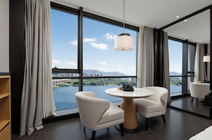 Habitación de hotel moderna con un ventanal grande, dos sillas blancas, una mesa redonda y una lámpara colgante.