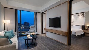 Una habitación de hotel moderna con un ventanal grande, un televisor de pantalla plana, un sofá, una silla, una mesita con flores y una cama.
