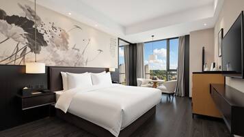 A modern hotel room with a large bed, a desk, a chair, a TV, and a view of the city.