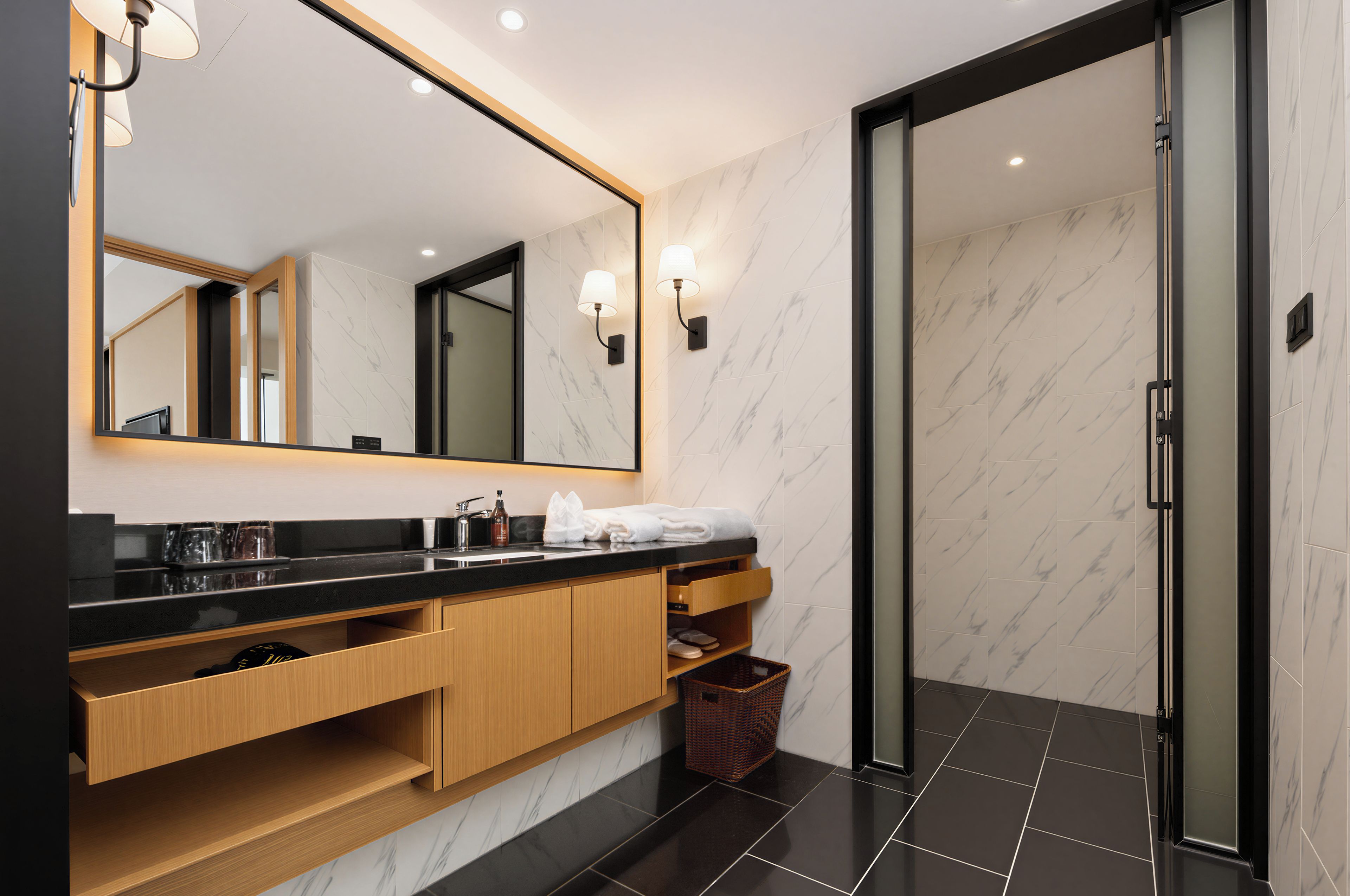 Une salle de bain moderne avec un grand miroir, un plan de toilette noir, des meubles en bois et une porte de douche en verre.