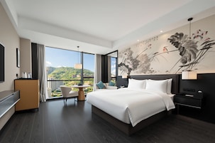 A modern hotel room with a large bed, a desk, a chair, and a view of a cityscape through the window.