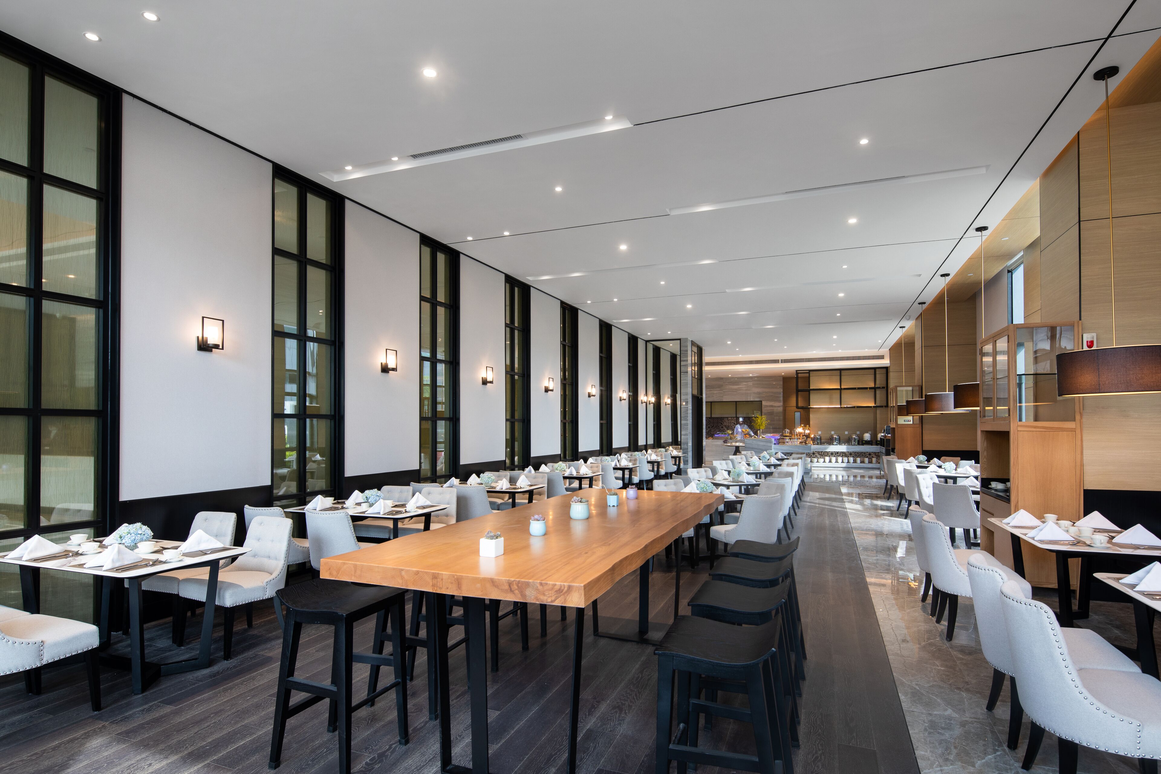Un'ampia sala da pranzo con lunghi tavoli e sedie, grandi finestre e moderne lampade.