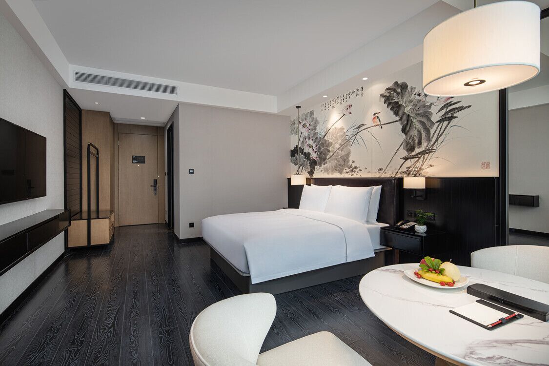A modern hotel room with a bed, bedside table, and a dining area with a table and chairs.