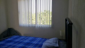 Una habitación pequeña y escasamente amoblada con una cama individual, una ventana con persianas verticales y una mesita de noche con lámpara.