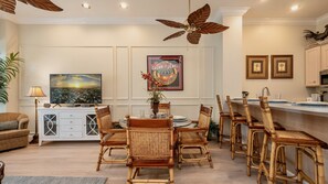 Un coin repas avec une table dressée pour six personnes, entouré de chaises en osier, d’un ventilateur de plafond et d’un téléviseur diffusant une scène de coucher de soleil.