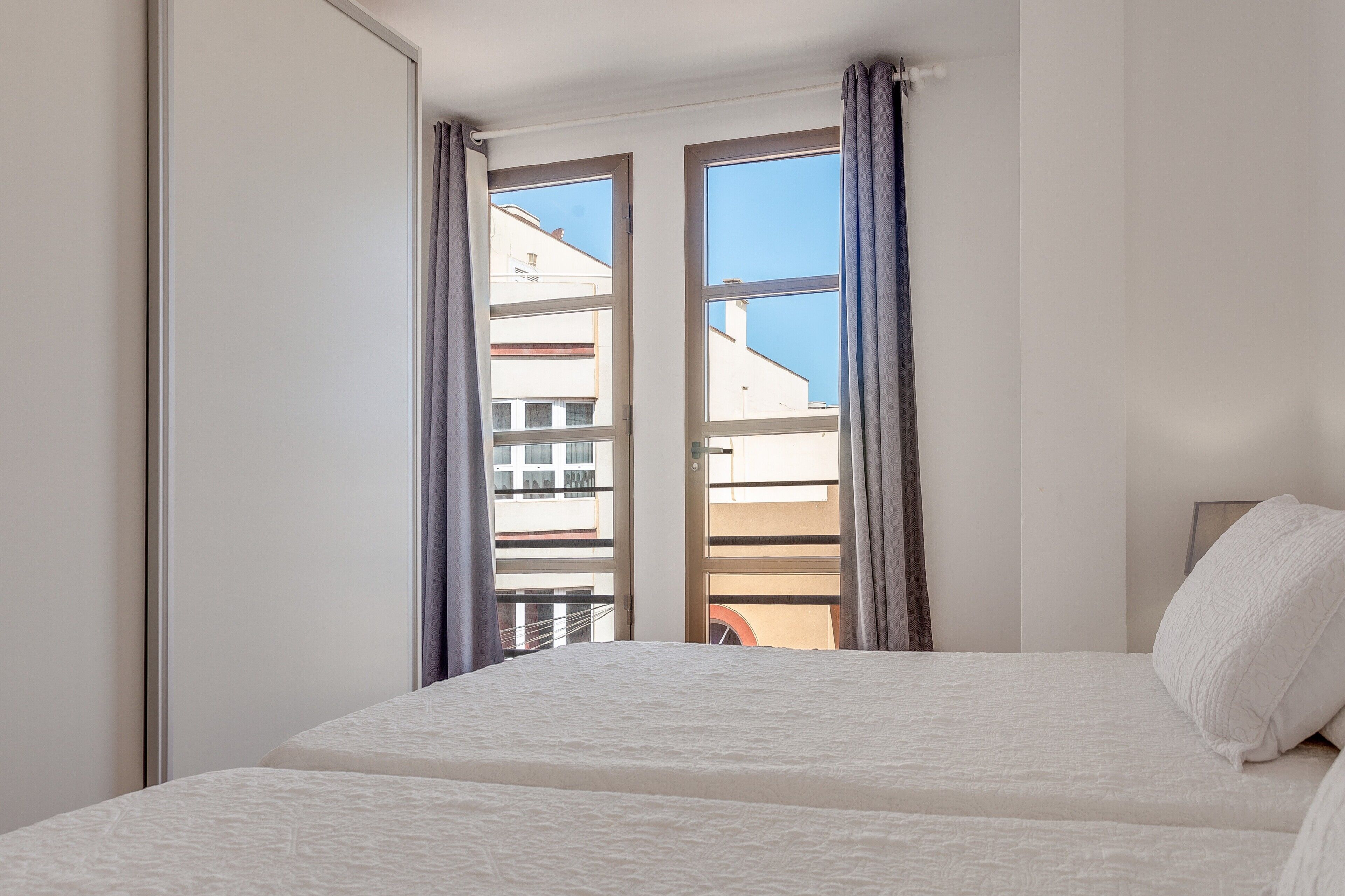 A bedroom with a bed, a window with curtains, and a view of buildings outside.