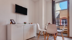Una habitación moderna con televisor de pantalla plana montado en la pared, un mueble blanco con cajones, una mesa de comedor con sillas blancas y un amplio ventanal con vistas a un edificio.