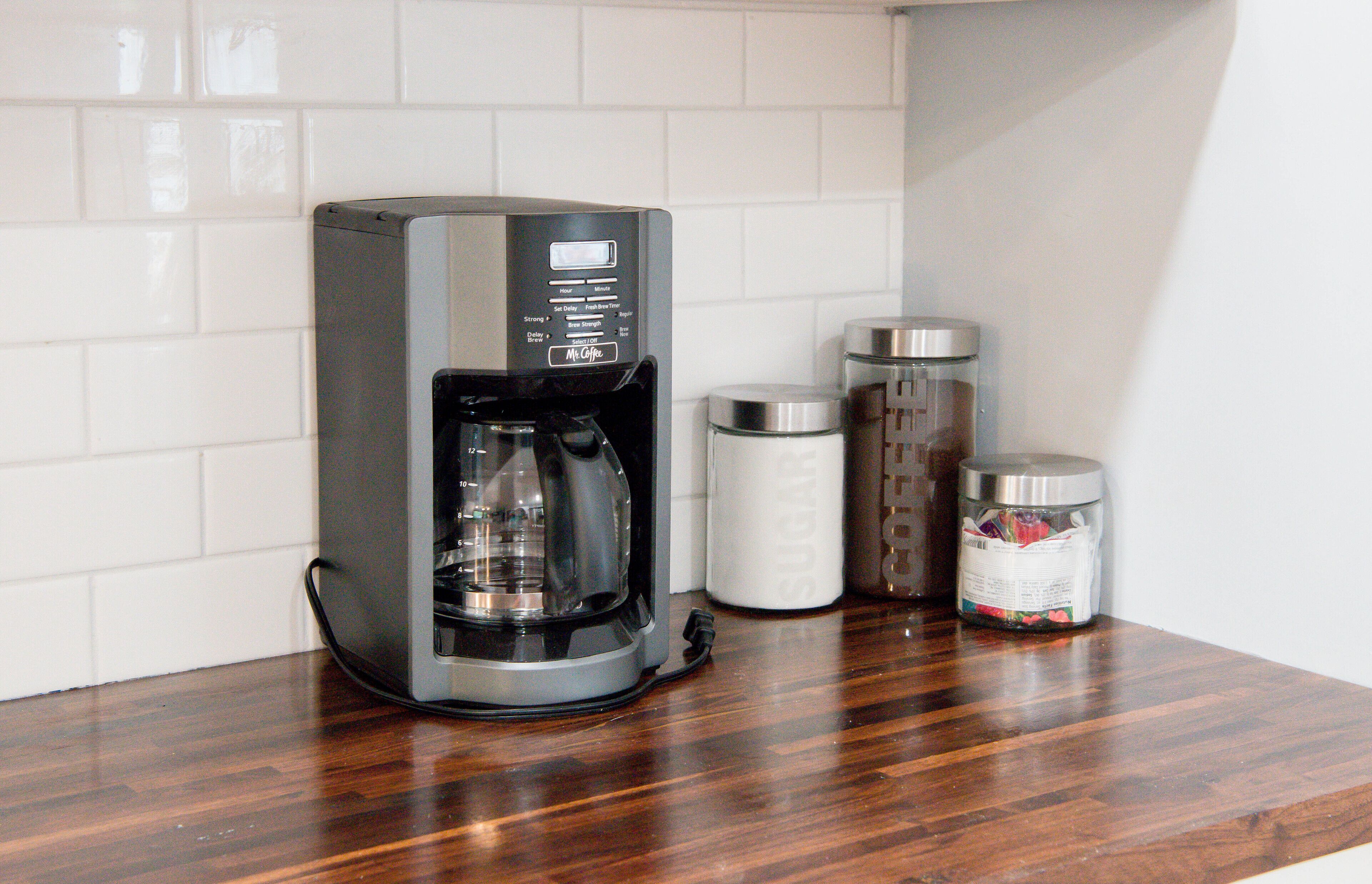 A coffee maker on a wooden countertop with white tiled walls and stainless steel containers.