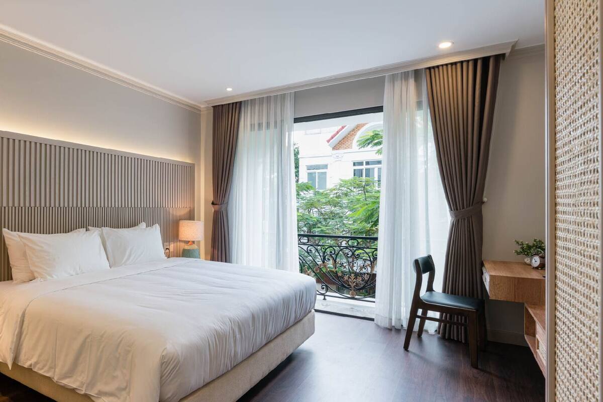 A hotel room with a large bed, a nightstand, a chair, a small table with a lamp, and a view of greenery and buildings through the window.