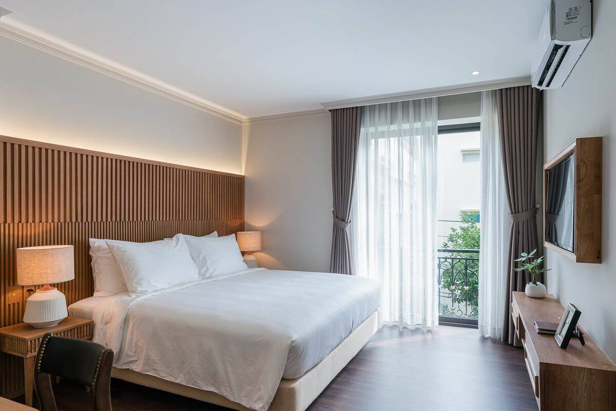 A hotel room with a large bed, two bedside tables with lamps, a wooden headboard, a small wooden dresser, a TV mounted on the wall, and a balcony with a view.