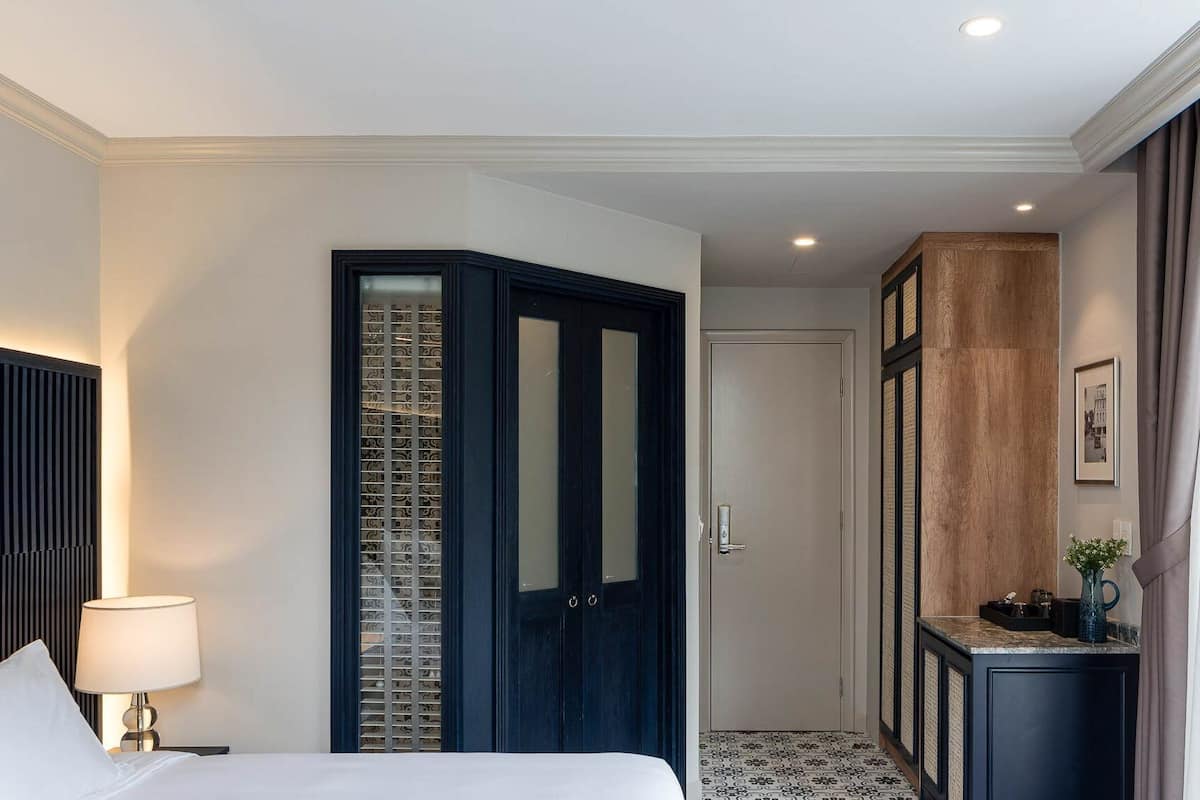 A hotel room with a bed, a nightstand, a lamp, a wardrobe, a door, a small cabinet, and a picture on the wall.