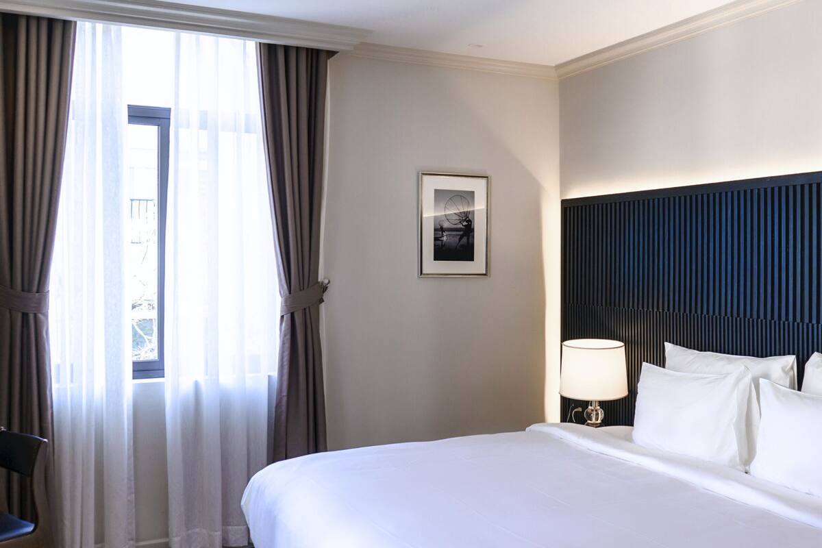 A hotel room with a bed, a nightstand with a lamp, a framed picture on the wall, and a window with curtains.