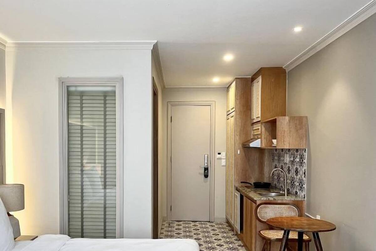 A compact hotel room with a bed, bedside lamp, and a small kitchenette with a sink and bar stools.