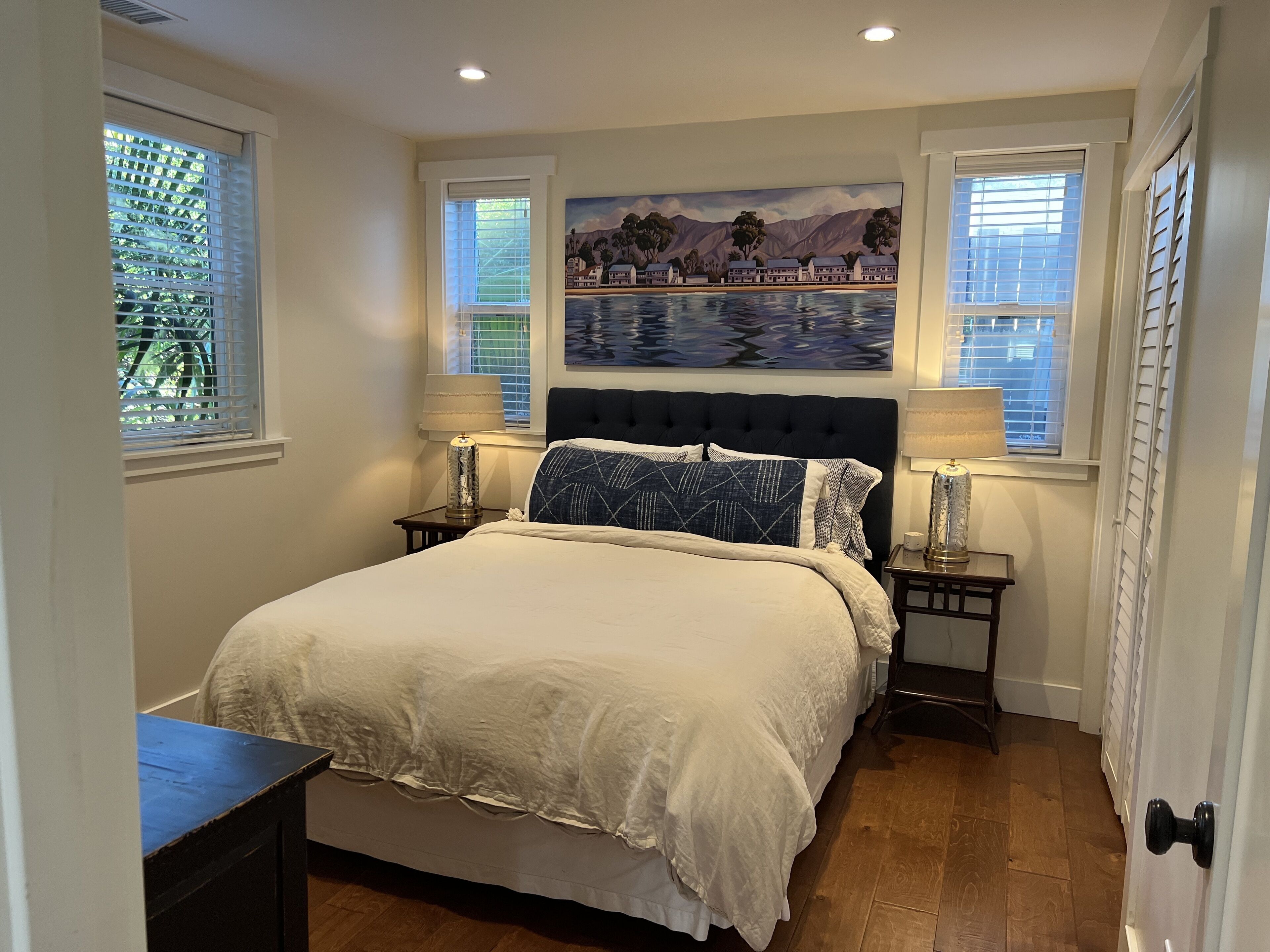 A bedroom with a bed, two nightstands, a painting above the bed, and windows with blinds.