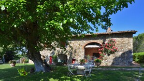 Un bâtiment en pierre avec un grand arbre devant, une table de patio et des chaises.