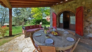 A stone table with plates, glasses, and a fruit bowl set outdoors under a wooden pergola.