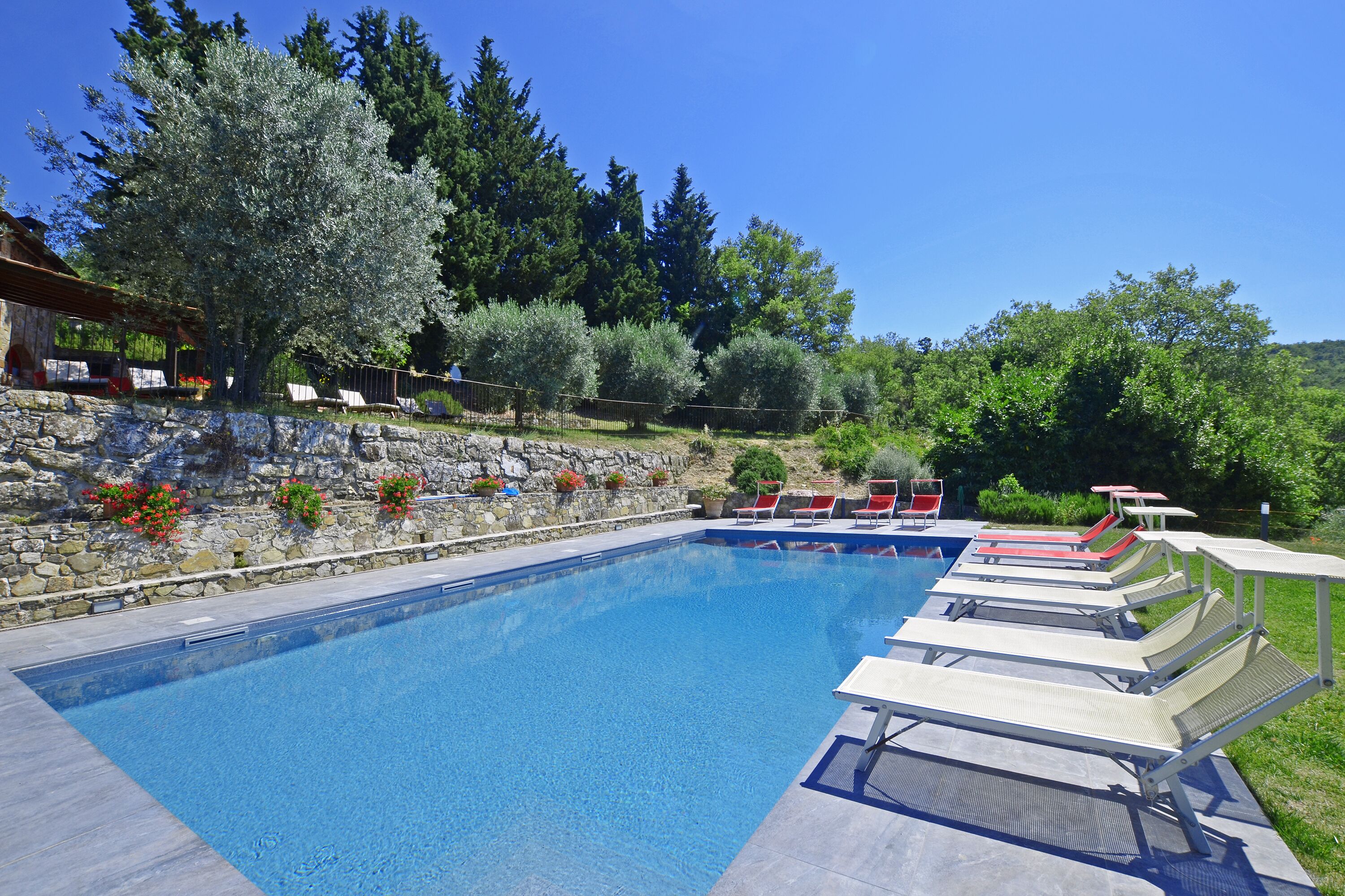 A swimming pool with lounge chairs, a stone wall, and trees in the background.