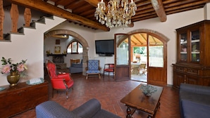A spacious living room with a wooden ceiling, a chandelier, and a television. There are multiple seating areas with chairs and sofas.