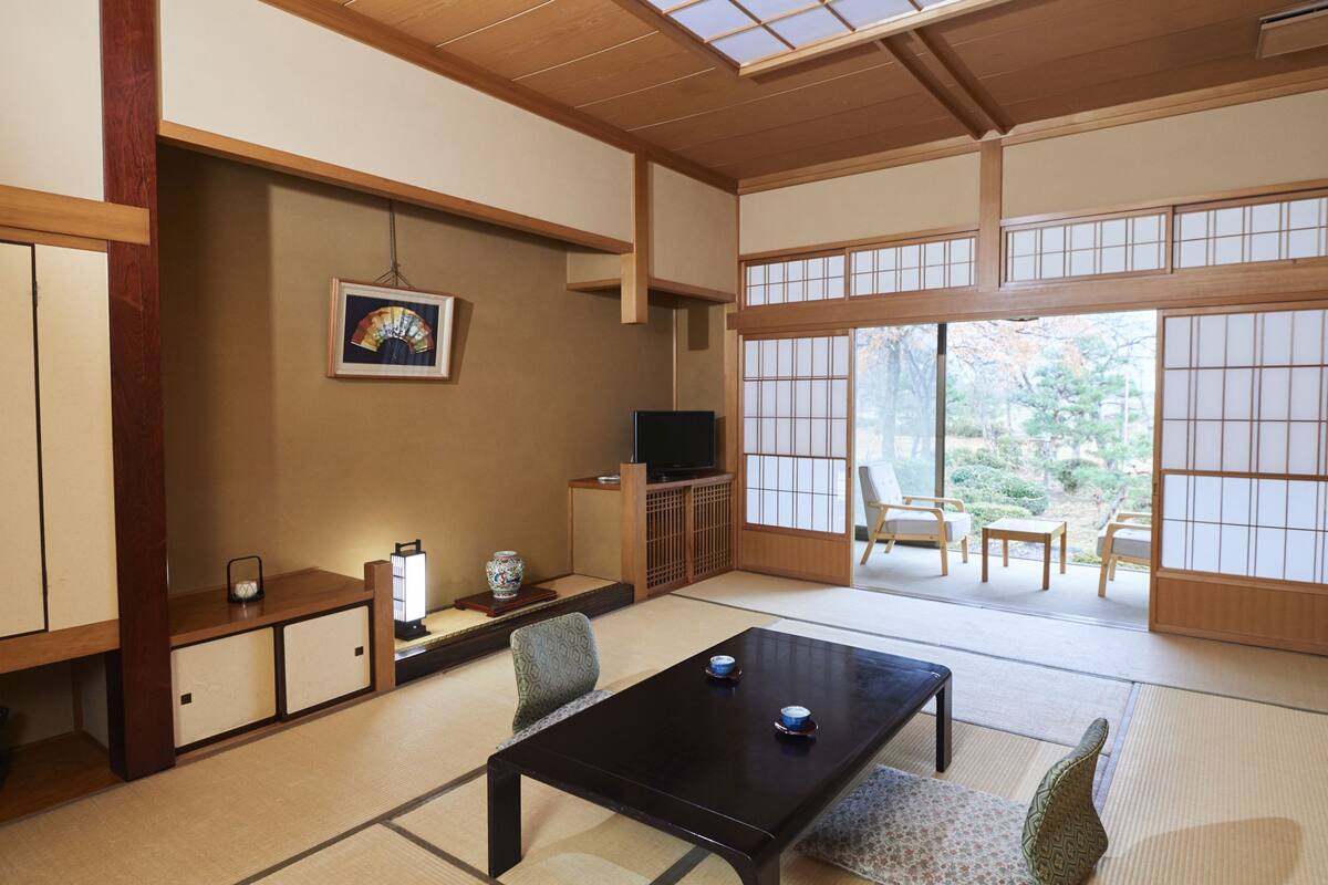 Una habitación japonesa tradicional con suelo de tatami, una mesa baja, sillas, un televisor y una puerta corrediza que da a un área exterior.