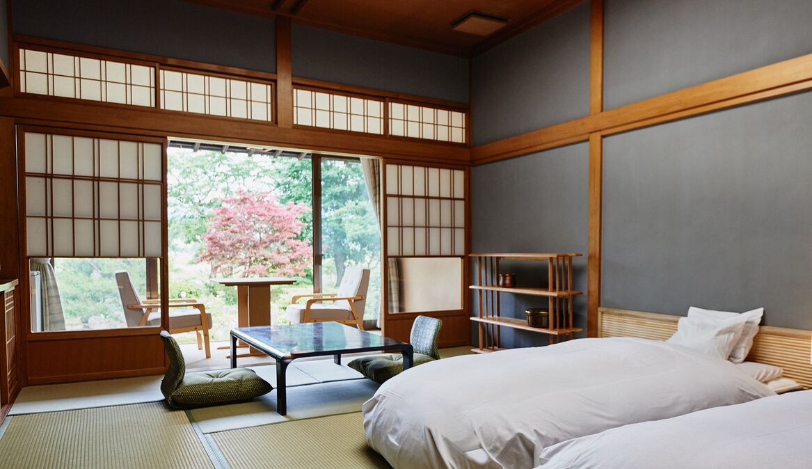 Una habitación tradicional japonesa con suelo de tatami, una mesa baja, sillas, una cama y vistas a flores de cerezo.