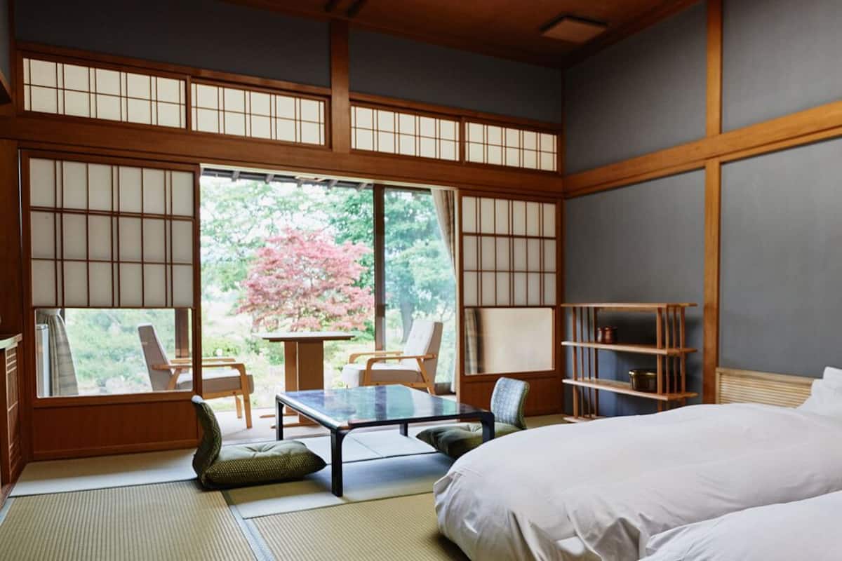 Una habitación tradicional japonesa con suelo de tatami, una mesa baja, sillas, una cama y vistas a flores de cerezo.