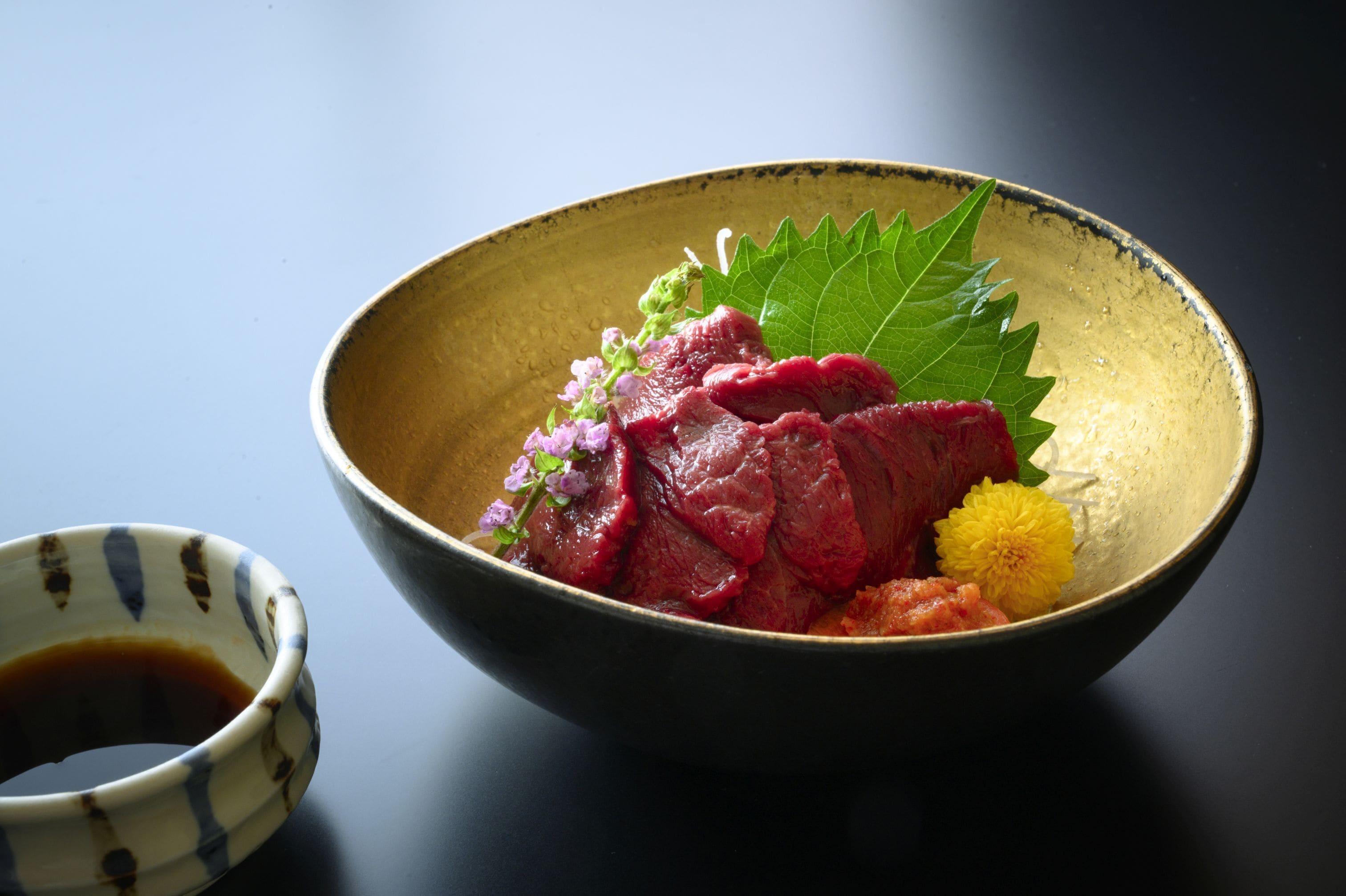 Eine Schüssel mit rohem Fleisch, einem Blatt und Blumen auf dunklem Untergrund.