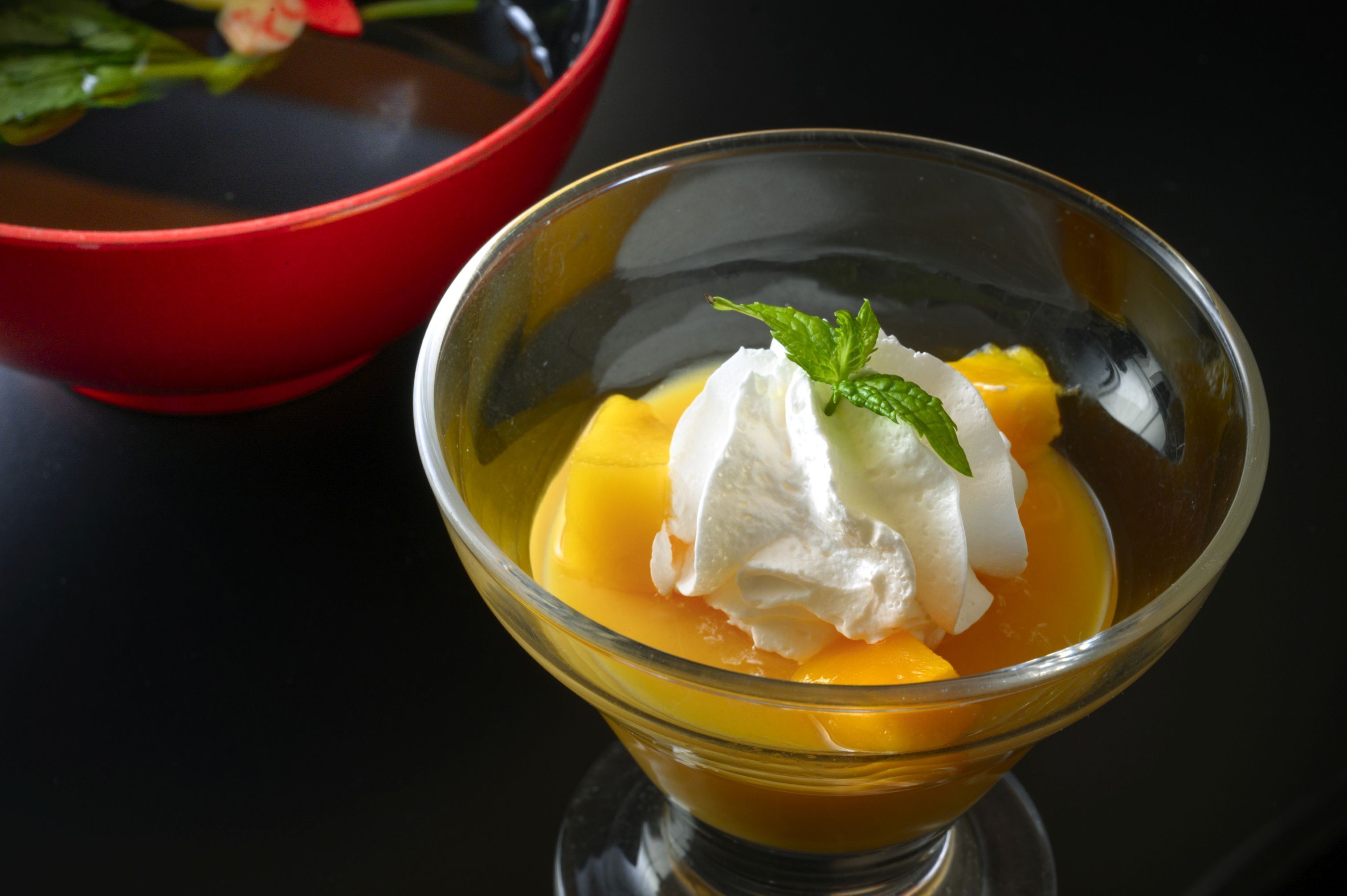 A dessert in a clear glass bowl with whipped cream and a mint leaf on top, accompanied by a red bowl in the background.
