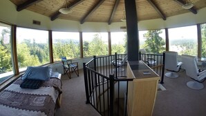 A spacious room with a high wooden ceiling, large windows offering a view of a forested landscape, a cozy seating area with a patterned rug, and a small bar counter. - Hilltop ocean view round house (Florence)