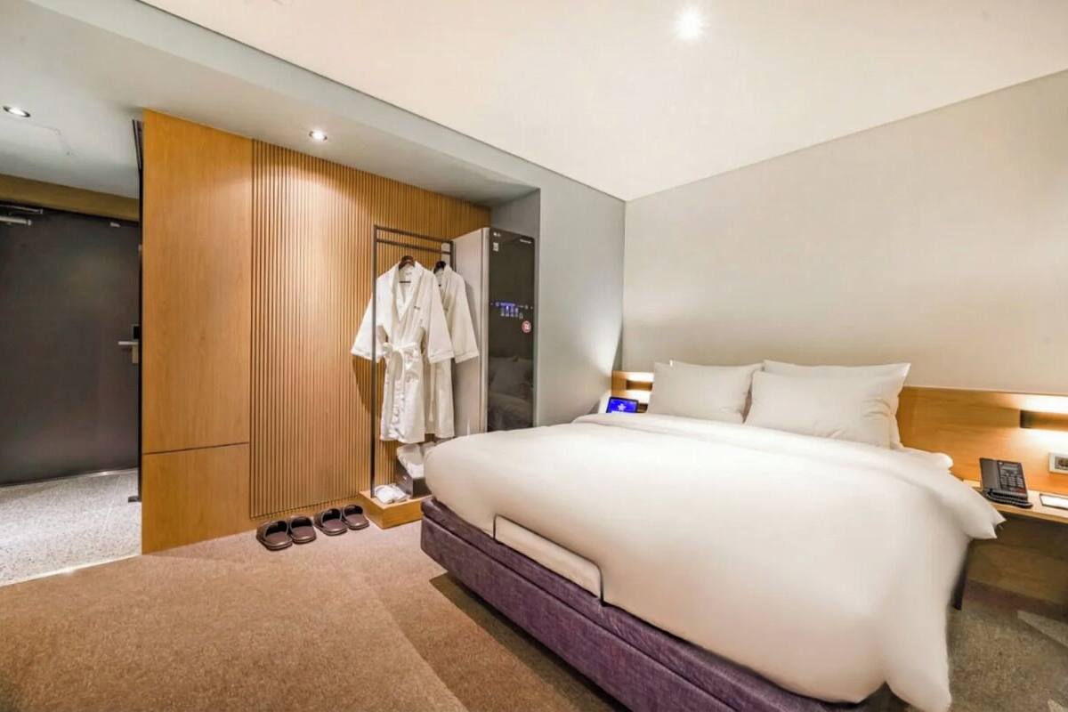 Habitación de hotel con una cama grande, un armario, un espejo, una mesita de noche con lámpara y un par de zapatillas en el suelo.