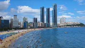 A beach with people and water activities, a city skyline with tall buildings, and a hill with buildings on top. - Haeundae Thevan Hotel (Busan)