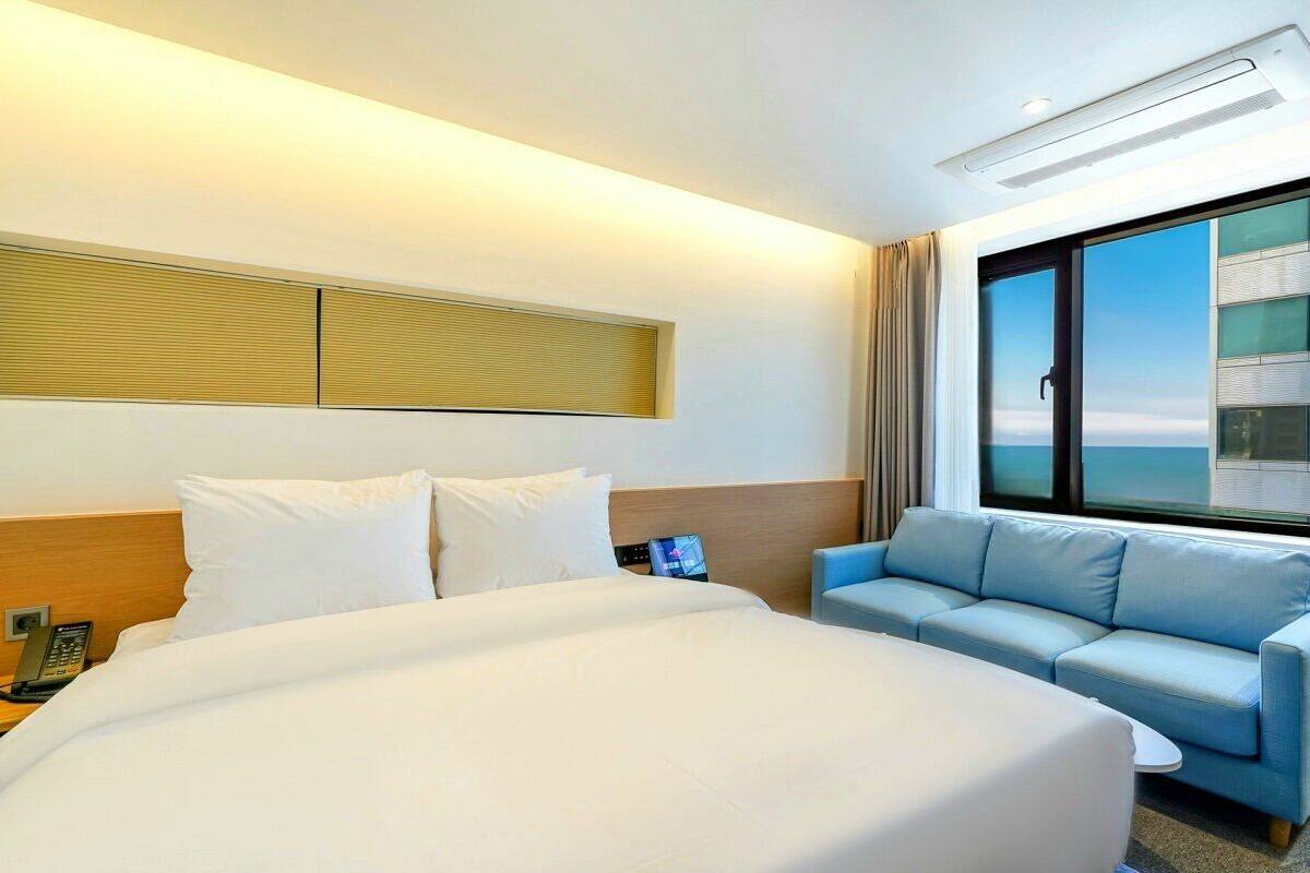 Habitación de hotel con una cama grande, un sofá, una mesita de noche con teléfono y una ventana con vistas a un edificio.