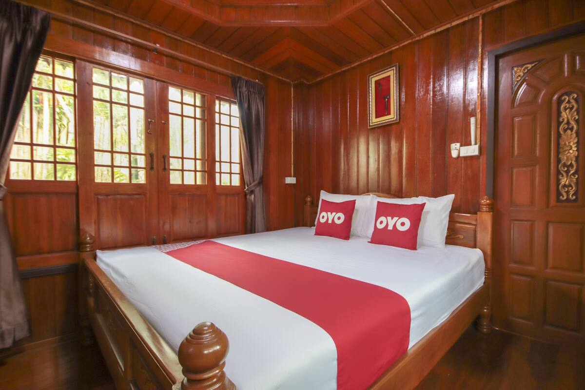 Uma cama de madeira com dossel, coberta por um edredom branco e vermelho, duas almofadas vermelhas com a inscrição "OYO" e uma cabeceira de madeira.