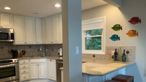 A kitchen with white cabinets, a microwave, an oven, a sink, and a window with a view of trees. There are fish wall decorations and a bar stool.