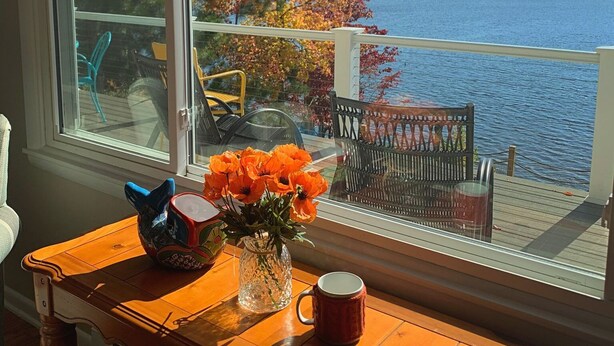 A wooden table with a vase of orange flowers and a mug, a chair, and a view of a lake with autumn trees.