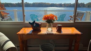 A wooden table with a vase of orange flowers, a ceramic fish bowl, and a mug. A window with a view of a lake and trees is in the background.