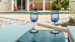 Due bicchieri di liquido blu su un tavolo di vetro, con una piscina e poltrone a sdraio sullo sfondo.