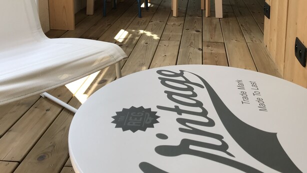Puinen terassi, jossa on valkoinen tuoli, pieni pöytä, jossa on logo, ja värikäs seinämaalaus.