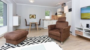 Een moderne woonkamer met een bruine bank, een voetenbankje en een kamerkleed met een patroon. Er is een eethoek met een tafel en stoelen, en een keuken op de achtergrond.