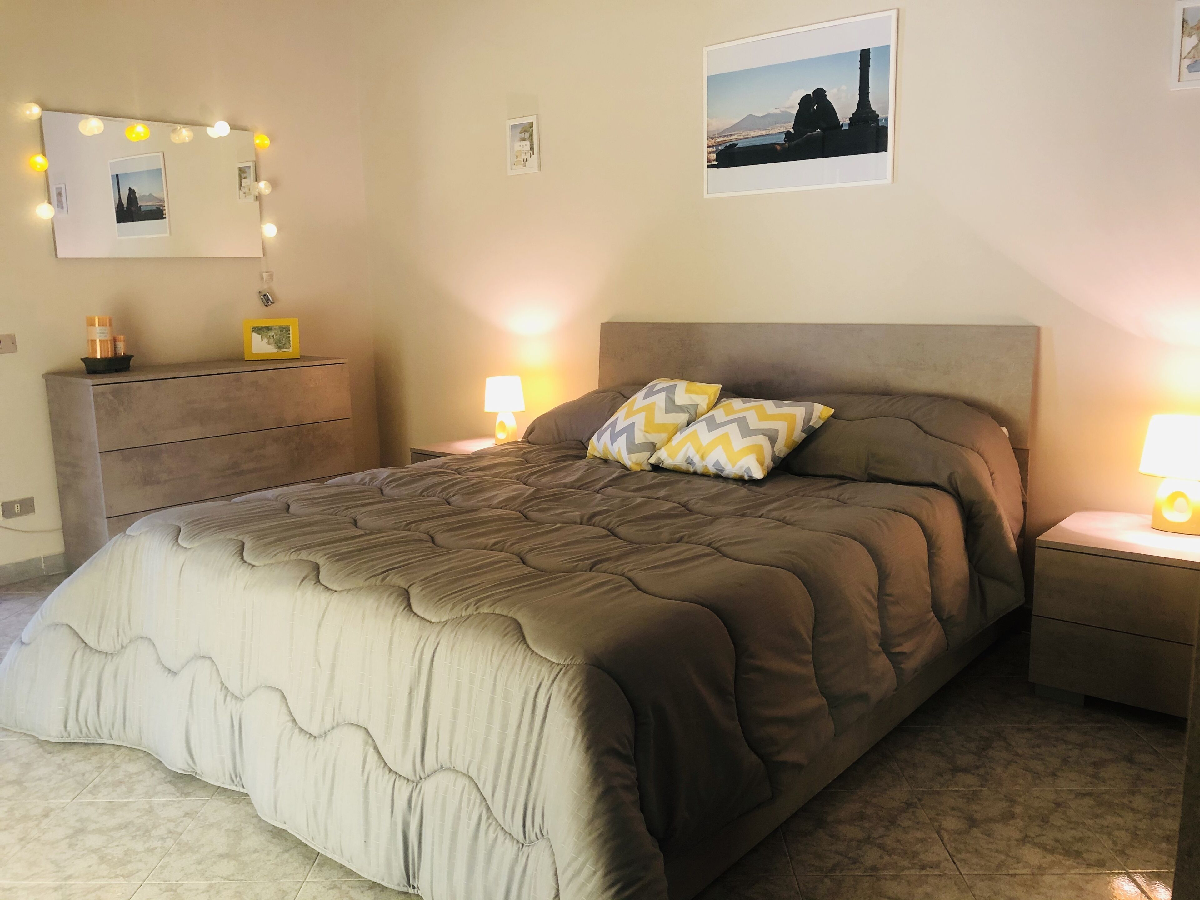 A bedroom with a large bed, a dresser, a nightstand with a lamp, and framed pictures on the wall.