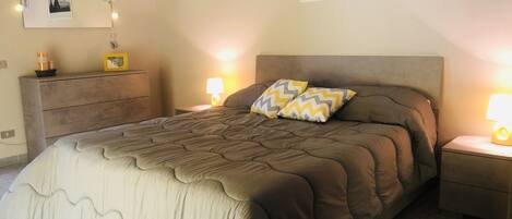 A bedroom with a large bed, a dresser, a nightstand with a lamp, and framed pictures on the wall.