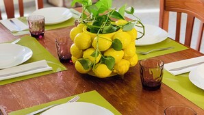 A dining table set with plates, cutlery, and glasses, with a centerpiece of lemons and greenery.