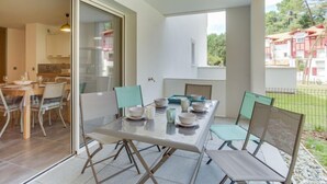 A dining area with a glass table and chairs, a kitchen visible through an open door, and a view of a residential area with houses and greenery. - Domaine Aïga by Interhome (Hossegor)
