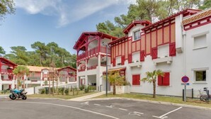 A row of white buildings with red accents, featuring balconies and a parking area with a bicycle symbol. - Domaine Aïga by Interhome (Hossegor)