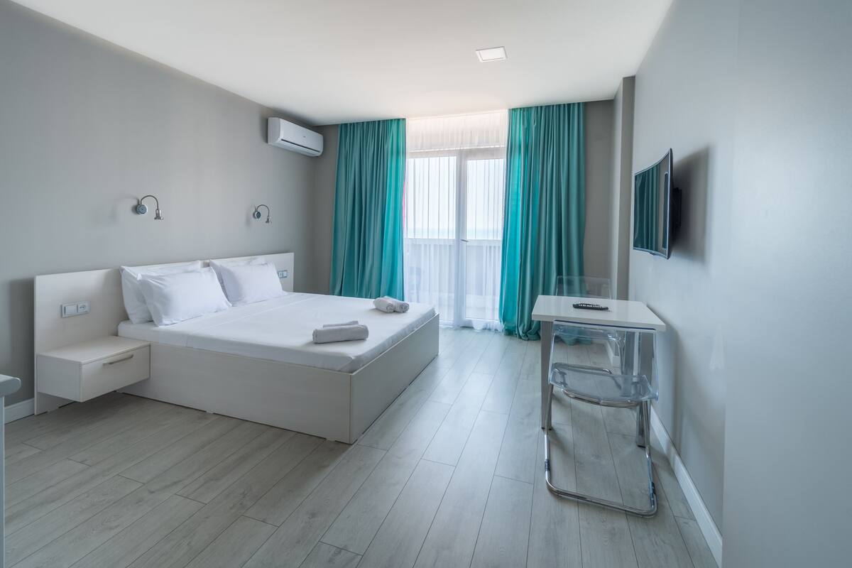 A modern hotel room with a large bed, a bedside table, a TV mounted on the wall, and a window with curtains.