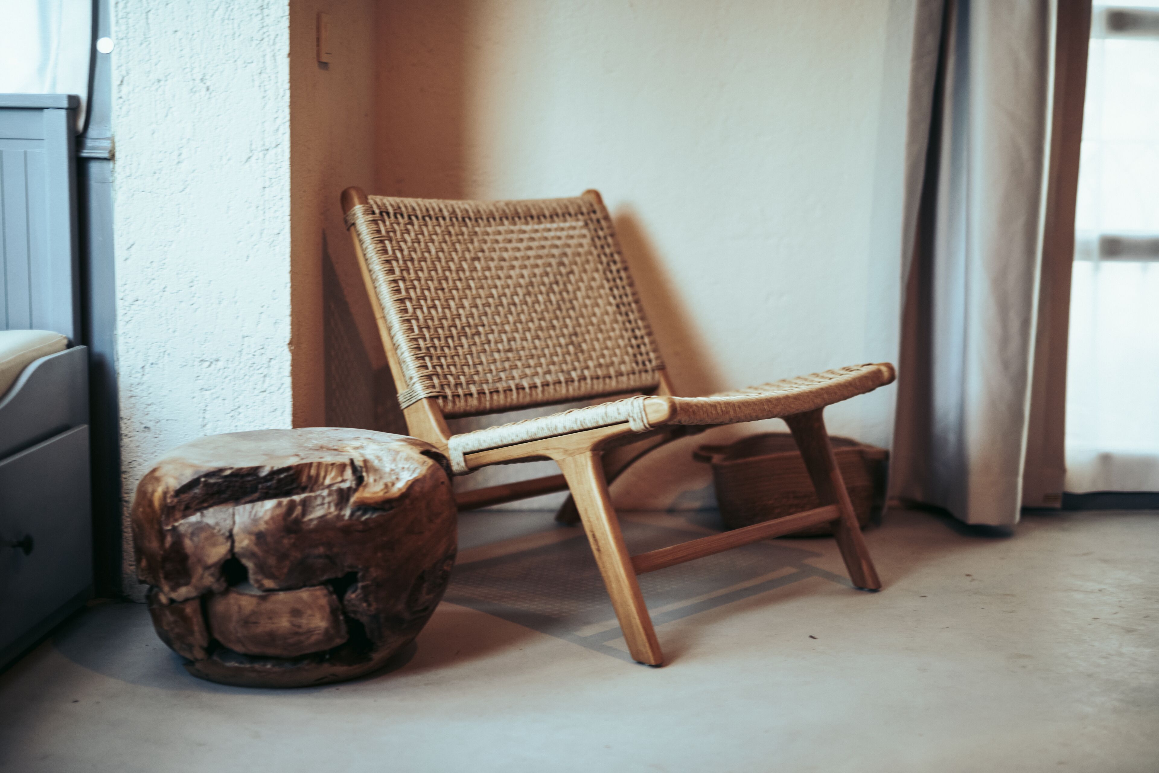 A wicker chair with a wooden base, a small round stool, and a white wall with a window.