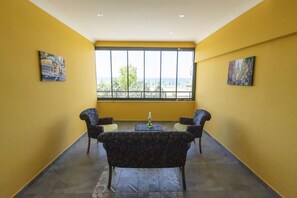 Una habitación con paredes amarillas, dos sillones, una mesita con un florero y ventanas amplias que dan vistas a árboles y edificios.