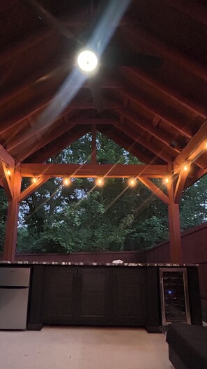 An outdoor covered area with a wooden ceiling, string lights, and a view of trees.