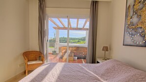 Un dormitorio con cama, una silla, una mesita de noche y vistas a un balcón con terraza de madera y vegetación.